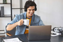 Junger Mann sitzt gut gelaunt mit einer Kassetasse in der Hand vor einem Laptop